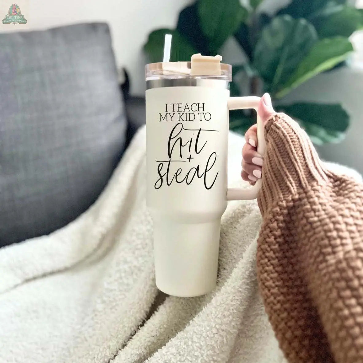 A person in a cozy sweater holds a 40oz Hit & Steal tumbler with a straw, featuring the phrase "I Teach My Kid to Hit + Steal." A soft blanket and a leafy green plant complete the comfy scene in the background.