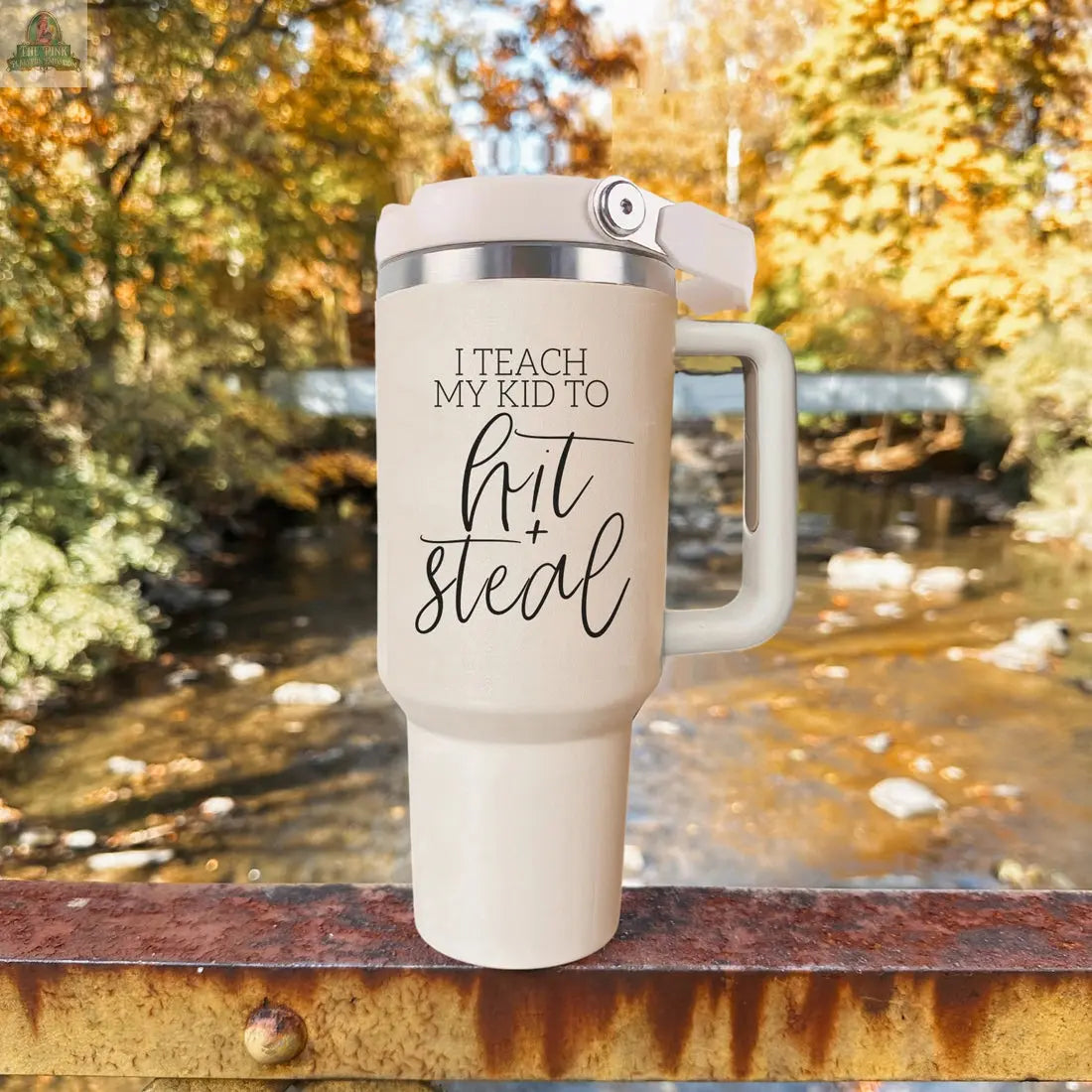 A 40oz beige Hit & Steal tumbler with "I Teach My Kid to Hit + Steal" rests on a rusty railing, overlooking a river and vibrant fall trees—a perfect accessory for baseball moms cheering at the game.