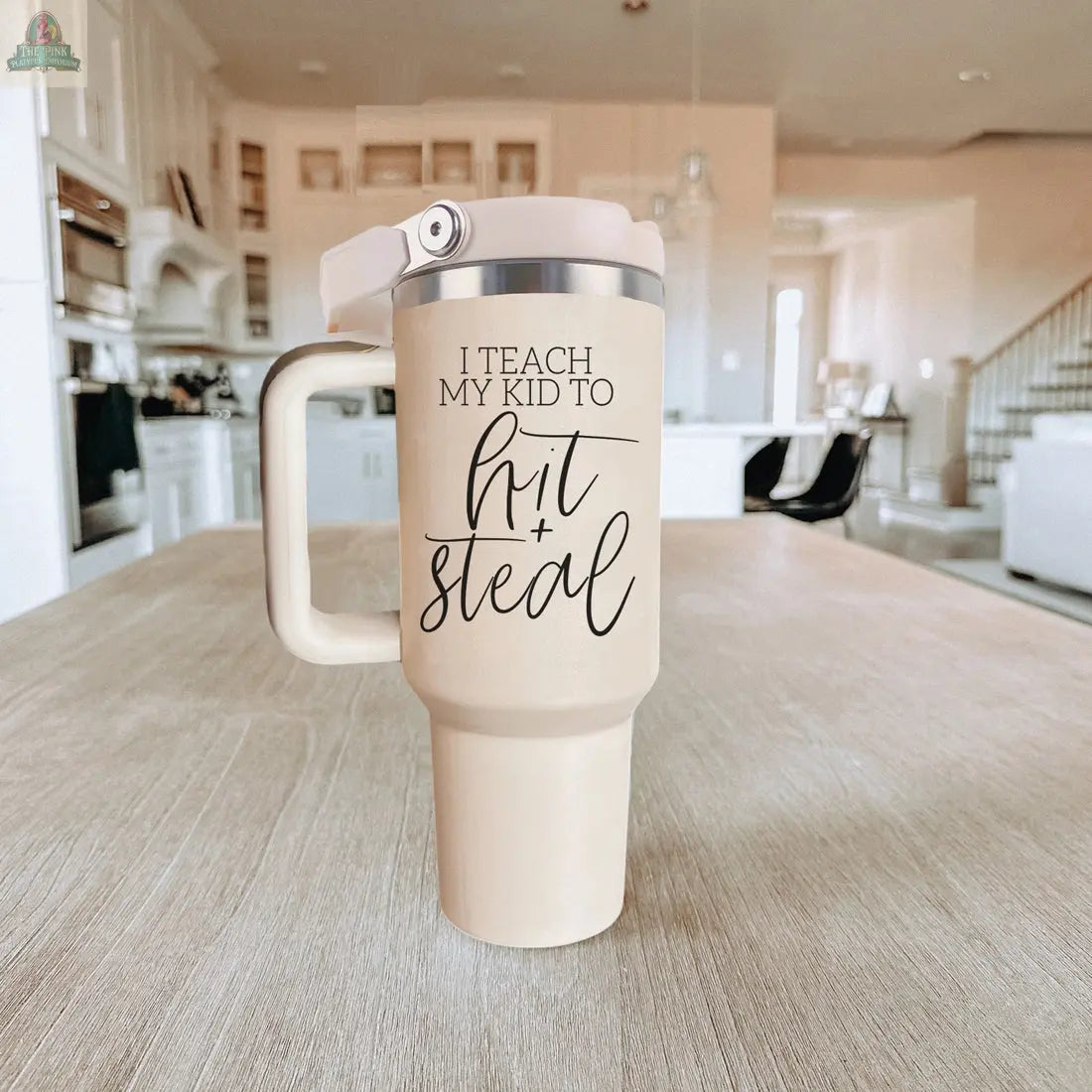 A beige 40oz Hit & Steal tumbler with a handle rests on a wooden table in a bright, modern kitchen. Its black script, “I Teach My Kid to Hit + Steal,” makes it the ideal baseball mom tumbler.