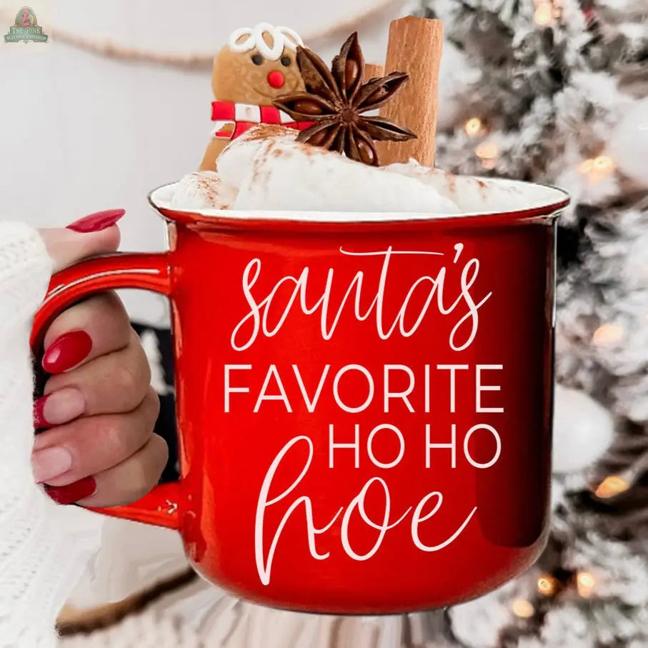 A hand with red-painted nails holds the Ho Ho Hoe Mug, filled with whipped cream, a gingerbread cookie, cinnamon stick, and star anise. A decorated tree is in the background.