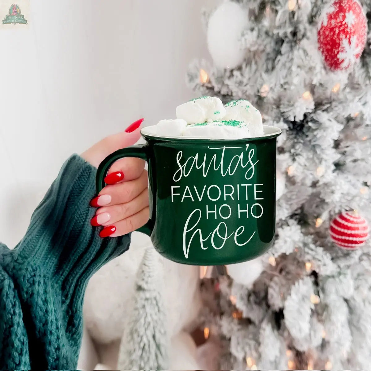 A hand with red and white nails holds the green Ho Ho Hoe Mug filled with marshmallows. “Santa’s Favorite Ho Ho Hoe” is written on the mug. A glowing white Christmas tree with ornaments shines in the background.