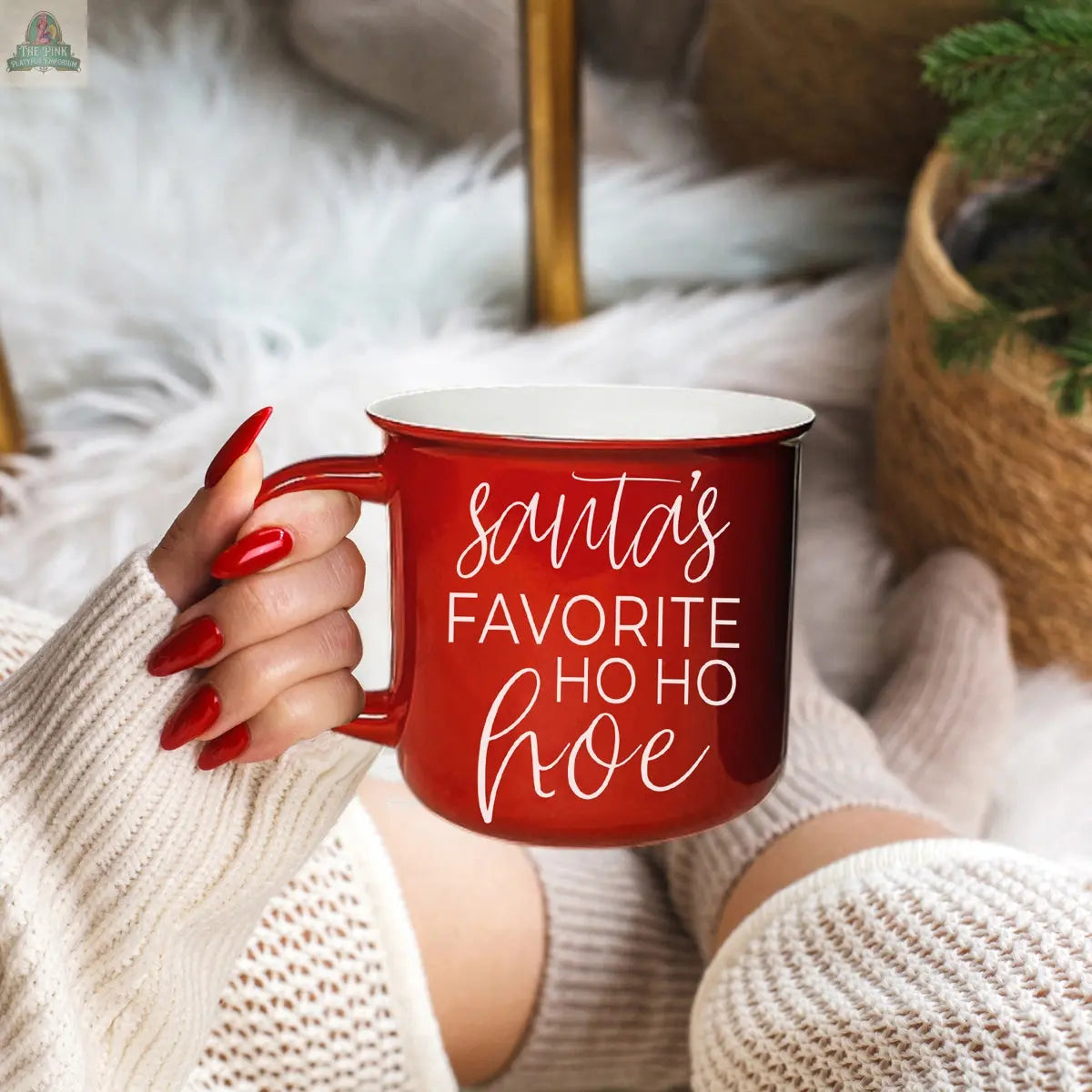 A person with red nails in a cream sweater holds the Ho Ho Hoe Mug, featuring "Santa's favorite ho ho hoe" text, while sitting on a white blanket beside a wicker basket and pine branches.