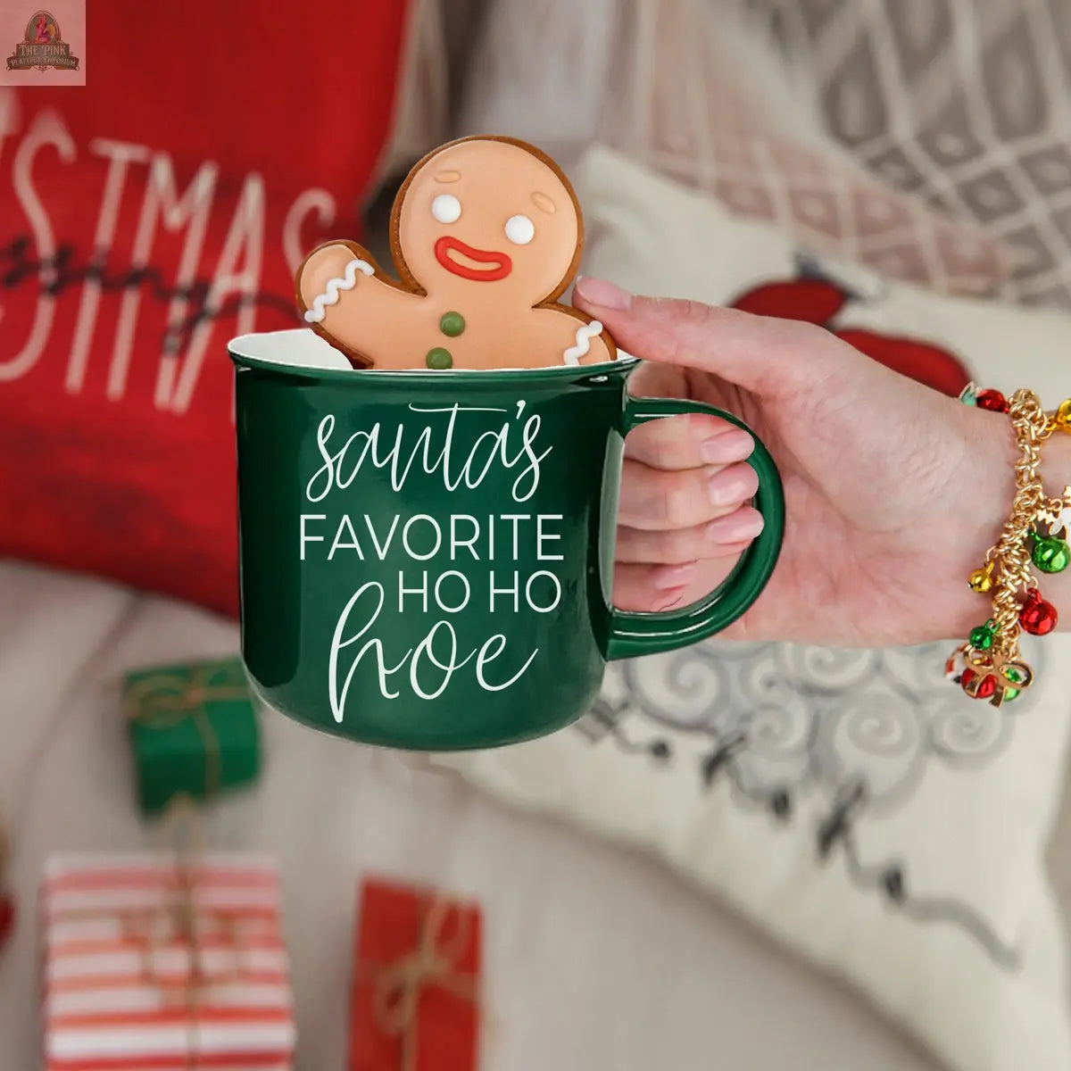A hand holds the green ceramic "Ho Ho Hoe Mug" with a smiling gingerbread man cookie inside, surrounded by blurred Christmas presents, a red pillow, and festive holiday decorations in the background.