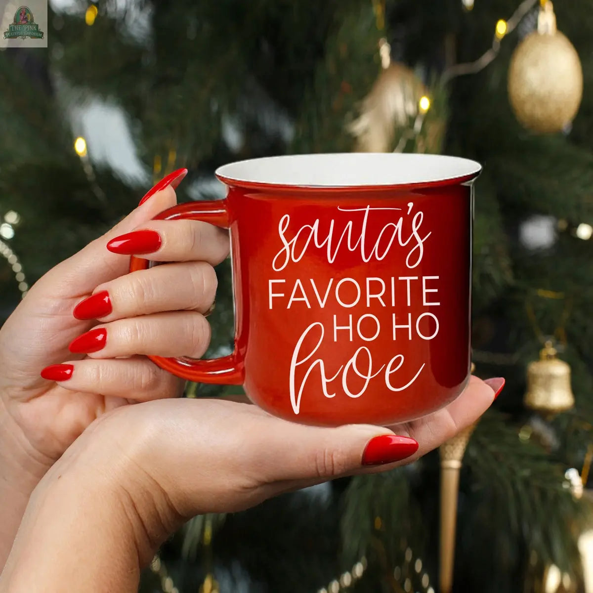 A person with long red nails holds the Ho Ho Hoe Mug, a handmade ceramic campfire mug featuring “Santa’s favorite ho ho hoe” in white script. A blurred Christmas tree with gold ornaments is in the background.