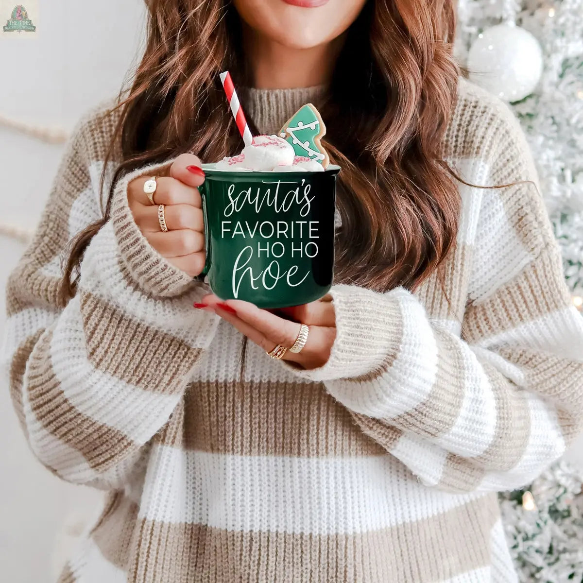 A woman in a brown and white striped sweater holds the Ho Ho Hoe Mug topped with whipped cream, a straw, and a gingerbread cookie. The festive mug is in front of a decorated Christmas tree.