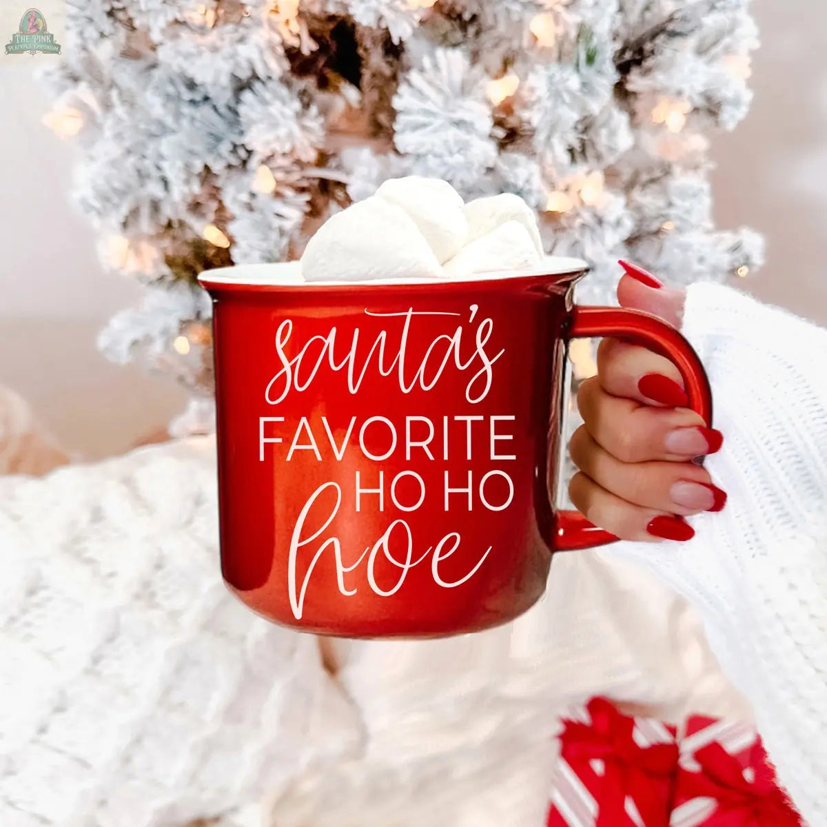A hand with red nail polish holds the Ho Ho Hoe Mug filled with marshmallows, featuring the phrase “Santa’s Favorite Ho Ho Hoe.” A white Christmas tree and wrapped gift appear in the background.