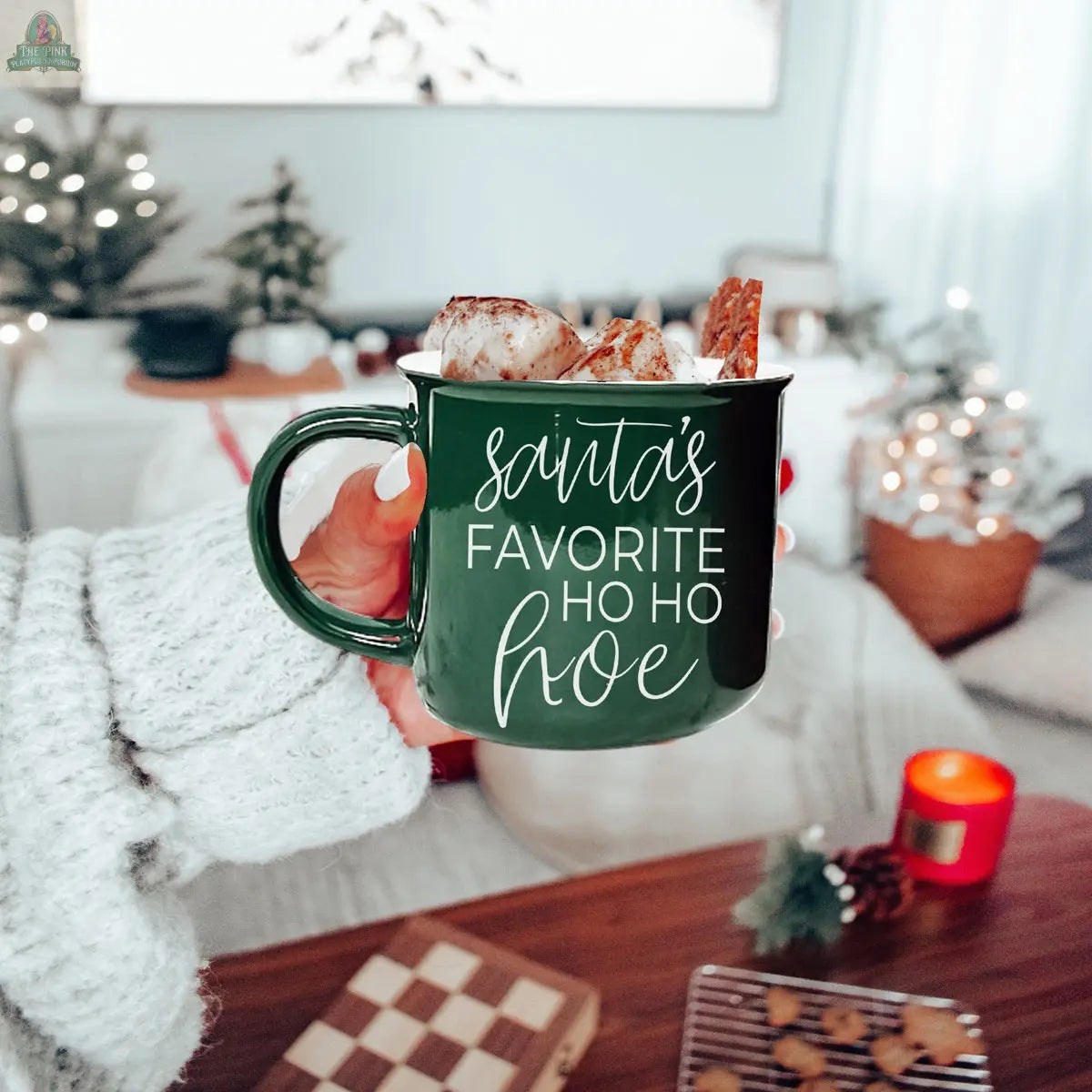 A hand in a cozy sweater holds the green ceramic Ho Ho Hoe Mug filled with hot chocolate, marshmallows, and a cinnamon stick. The festive living room in the background is decorated for Christmas.