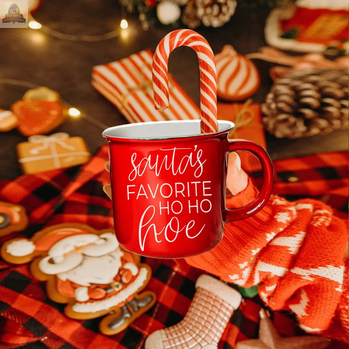 A gloved hand holds up the Ho Ho Hoe Mug, a red ceramic campfire mug with “Santa’s Favorite Ho Ho Hoe” on it and a candy cane inside, while holiday cookies, pinecones, and festive decorations are blurred in the background.