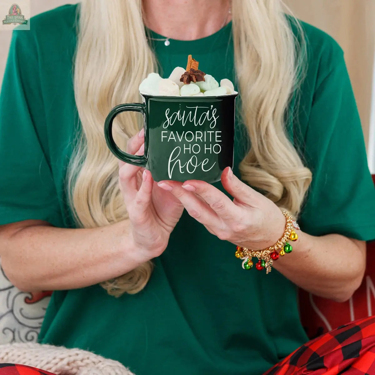 A person with long blonde hair, in a green shirt and festive bracelet, holds the Ho Ho Hoe Mug—a ceramic campfire-style mug topped with marshmallows and a cinnamon stick. The mug reads: Santa's favorite ho ho hoe.
