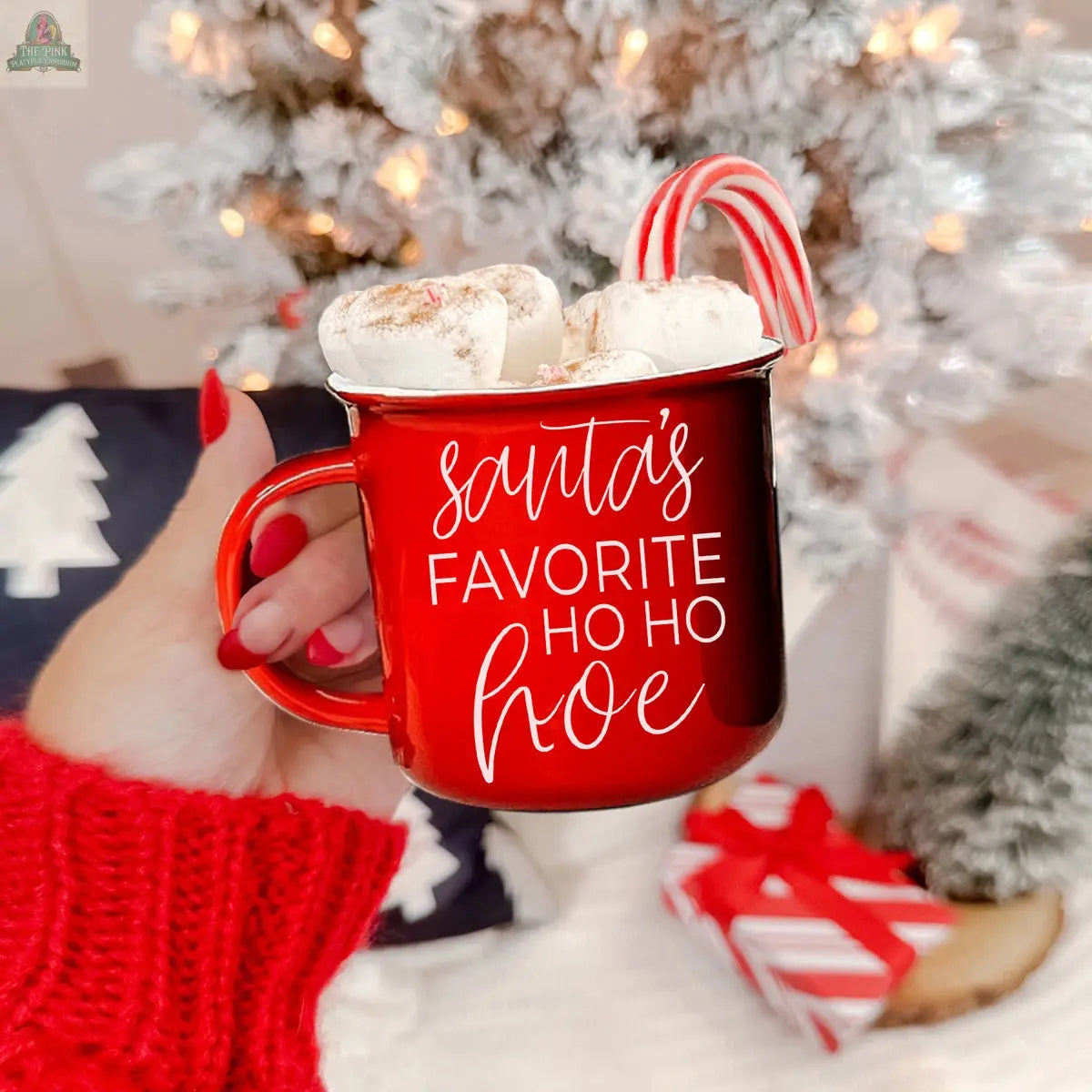 A hand in a red sweater holds the Ho Ho Hoe Mug, filled with hot chocolate, marshmallows, and a candy cane. The mug reads “Santa’s Favorite Ho Ho Hoe.” A white Christmas tree and wrapped gifts are in the background.