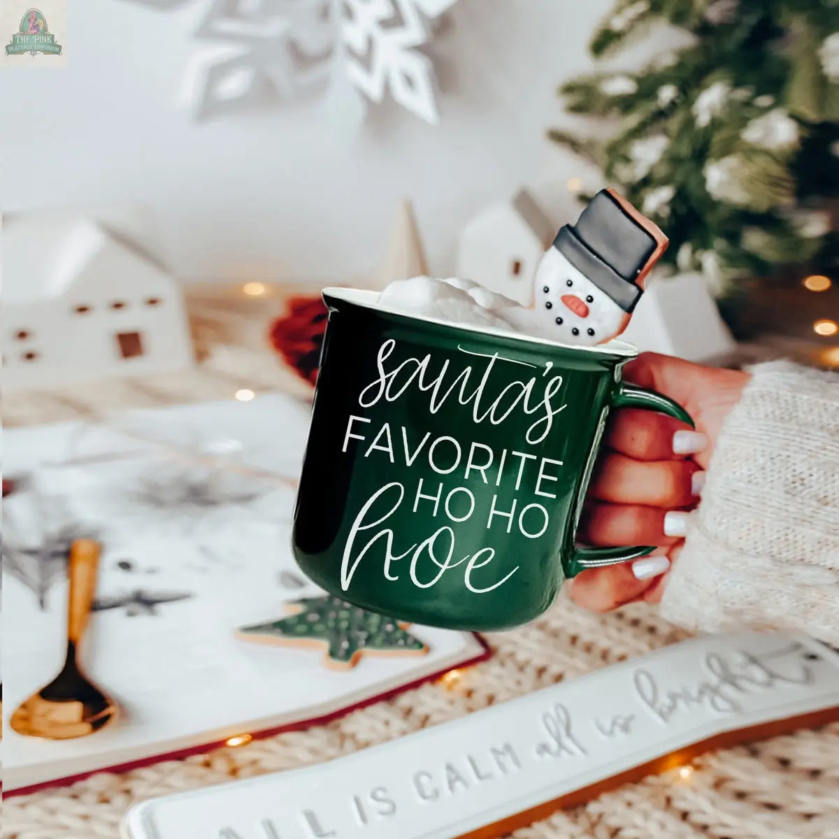 A hand holds the green ceramic Ho Ho Hoe Mug, filled with a hot drink, whipped cream, and topped with a snowman cookie. Festive holiday decorations blur in the background.