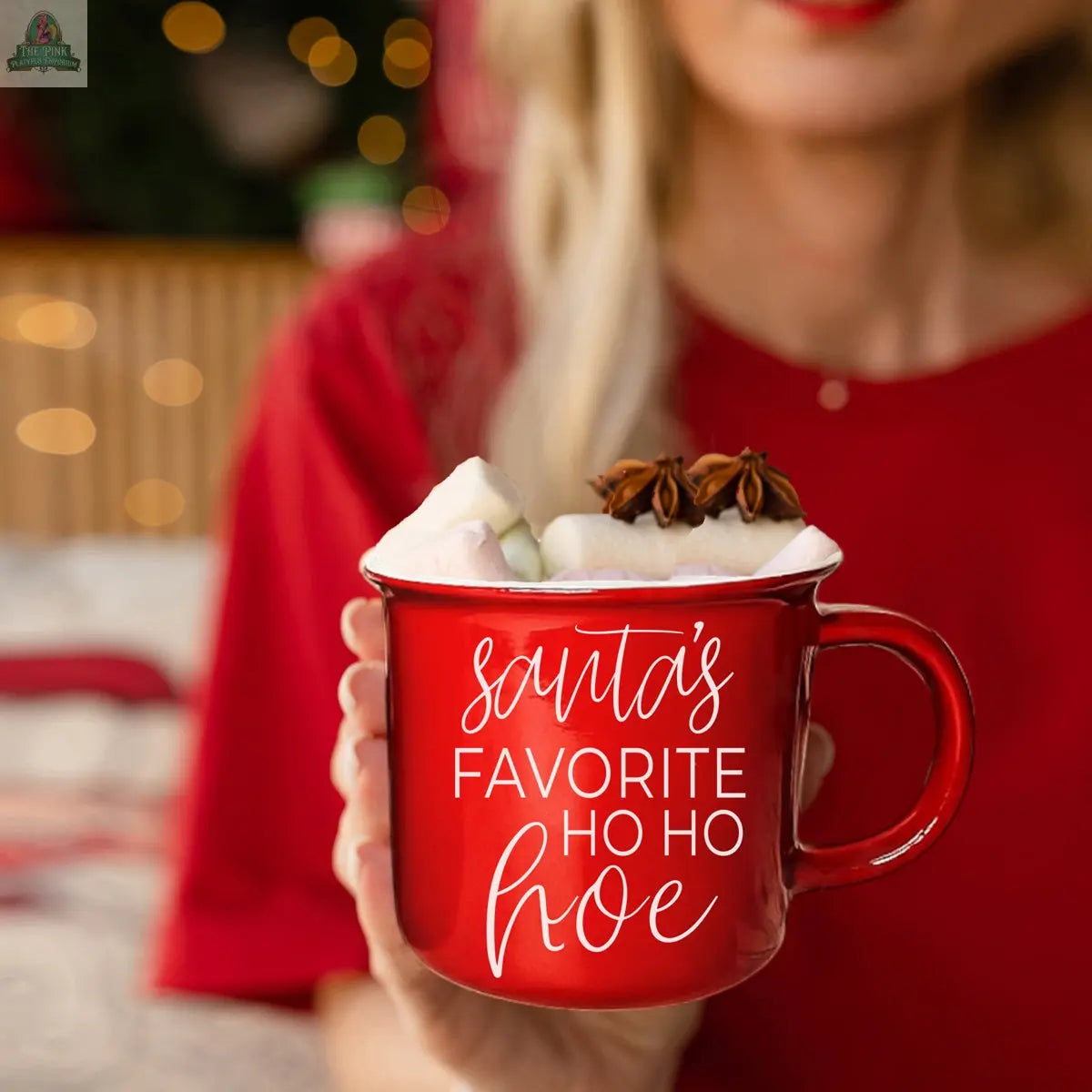 A woman in a red shirt holds the Ho Ho Hoe Mug, a festive ceramic campfire mug filled with marshmallows and star anise. “Santas favorite ho ho hoe” is written in white script. Soft holiday lights glow, highlighting the microwave-safe design.
