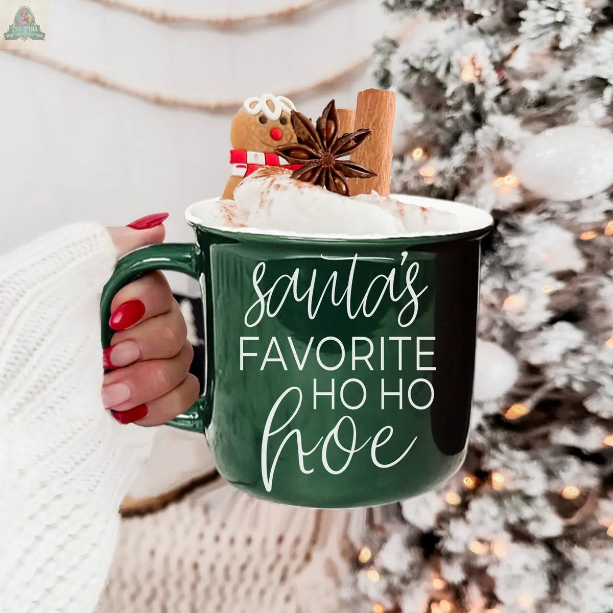 A hand with red-painted nails holds the green Ho Ho Hoe Mug filled with whipped cream, a cinnamon stick, a gingerbread cookie, and star anise, with a glowing decorated Christmas tree in the background.