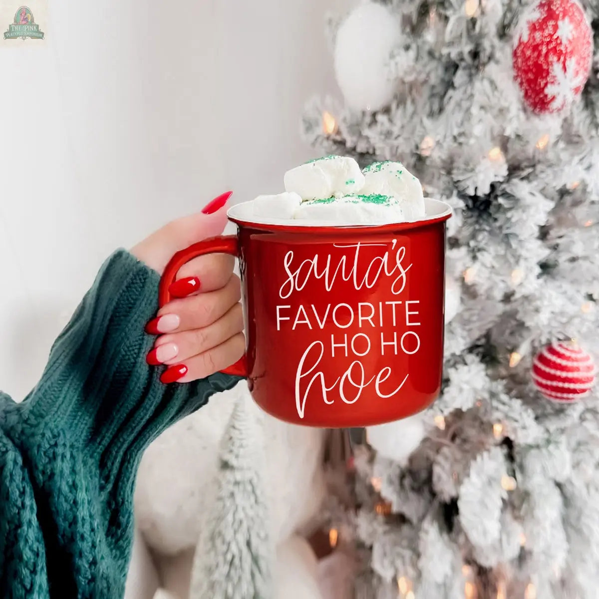 A hand with red nails holds the Ho Ho Hoe Mug, filled with marshmallows and featuring white text. A decorated white Christmas tree with red and white ornaments glows softly in the background.