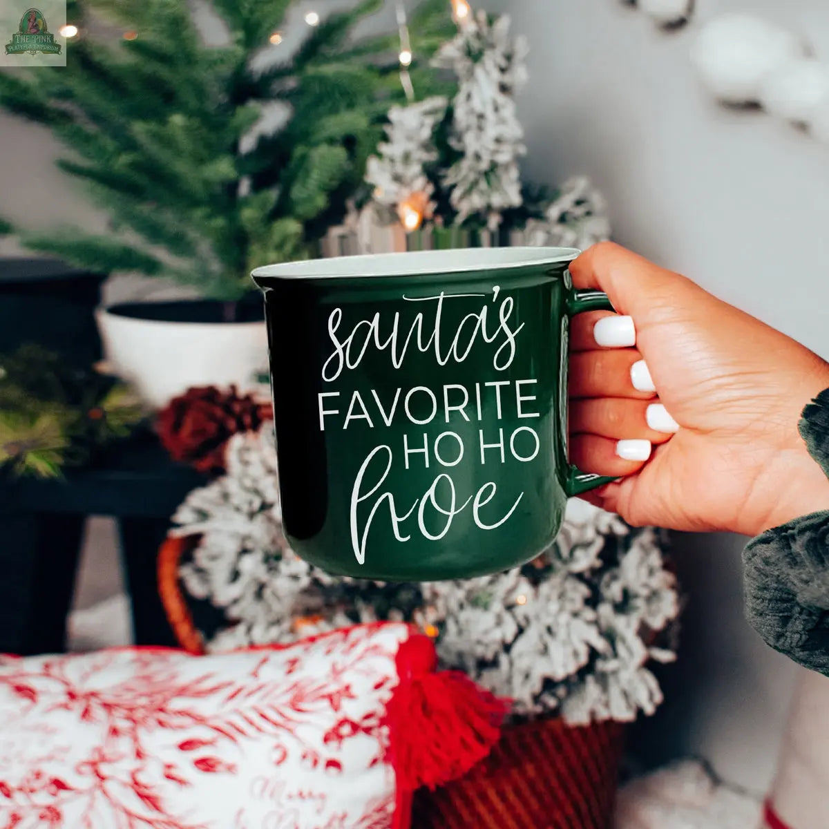 A hand with white nail polish holds the Ho Ho Hoe Mug, a green ceramic campfire-style mug with the phrase “Santa’s favorite ho ho hoe” in white script. A decorated Christmas tree and festive decor are blurred in the background.