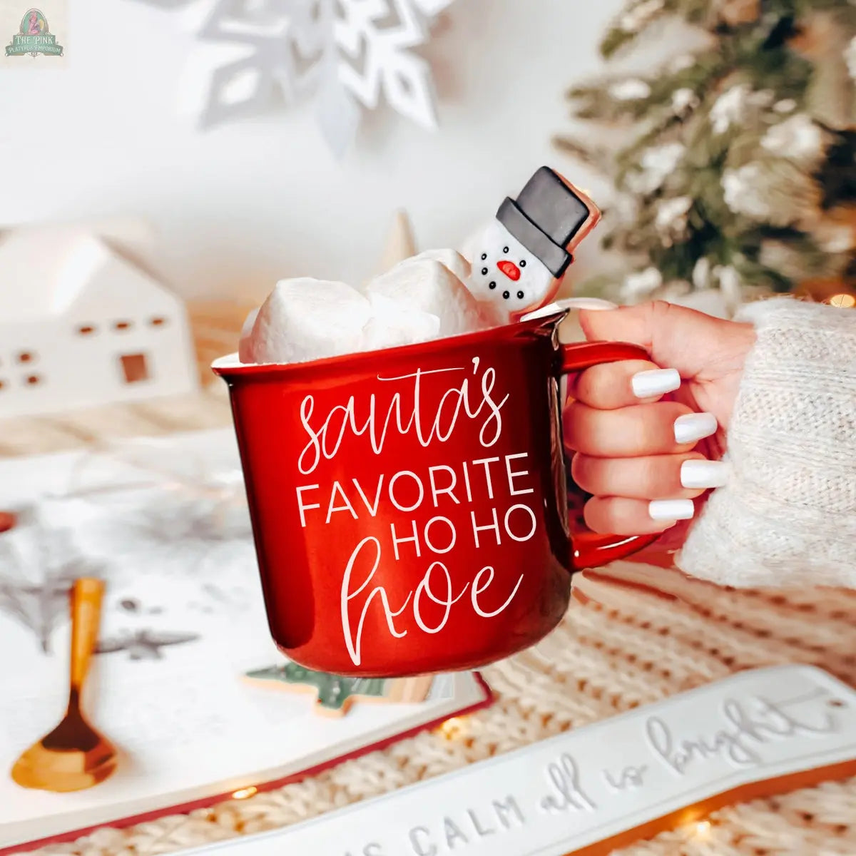 A hand with white manicured nails holds the red "Ho Ho Hoe Mug," filled with marshmallows and a snowman cookie. Holiday decor, a tree, and a book can be seen in the background.