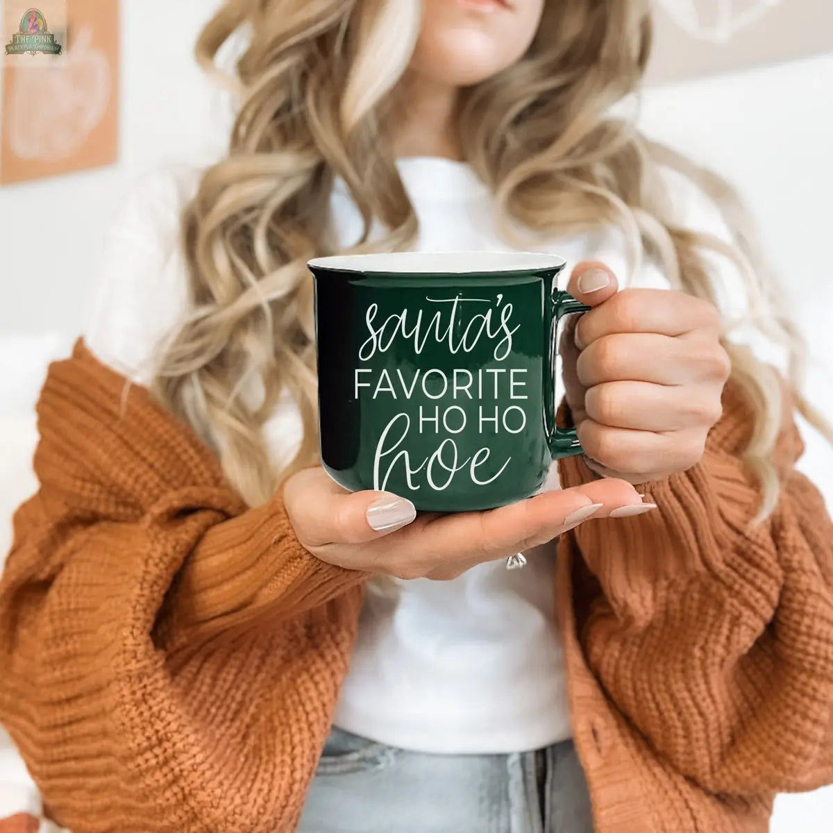 A woman with wavy blonde hair in a white shirt and orange cardigan holds the Ho Ho Hoe Mug, a green handmade ceramic campfire mug featuring "Santa's favorite ho ho hoe" in white script.