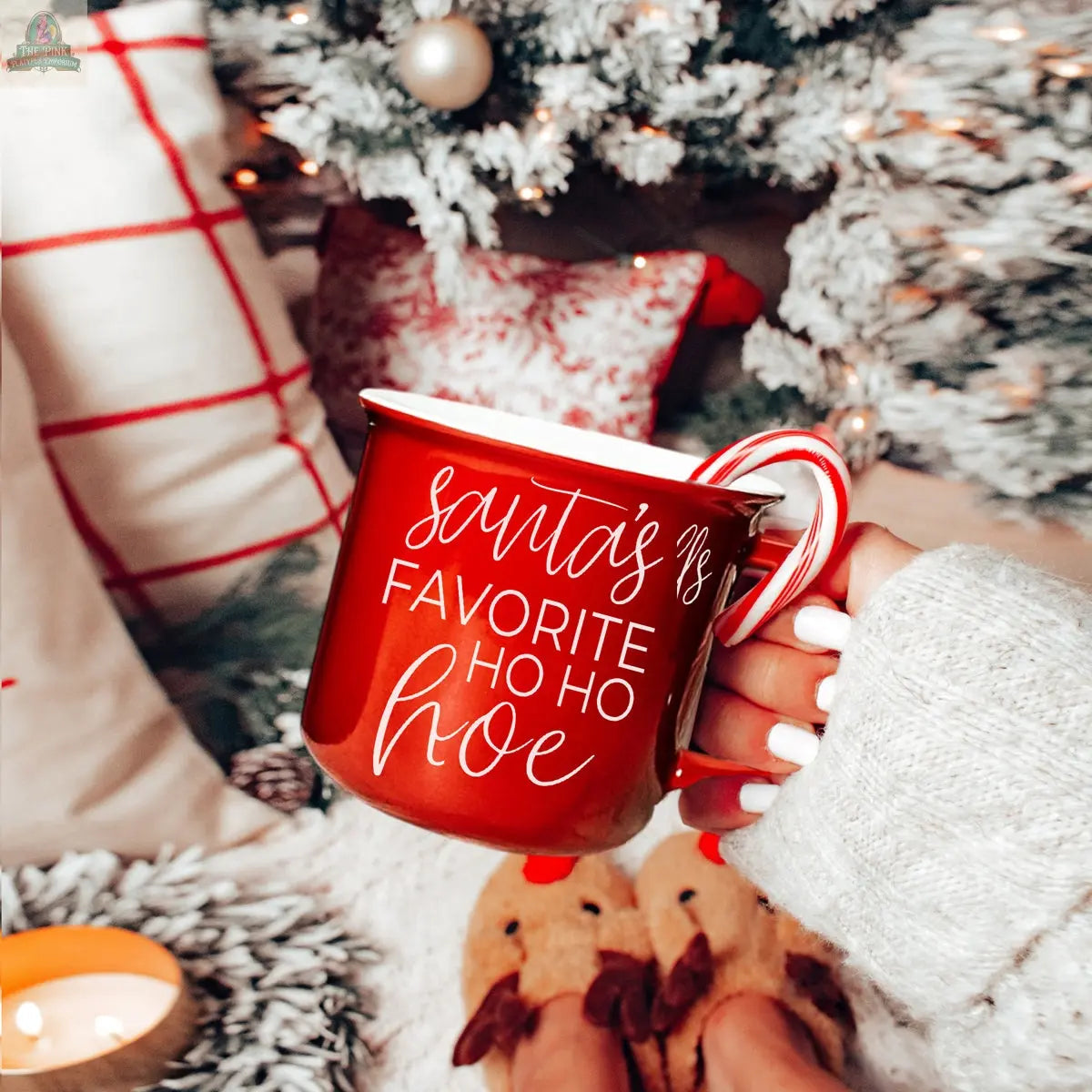 A hand holds the Ho Ho Hoe Mug, a red ceramic cup with white text, in front of a Christmas tree, cozy pillows, a lit candle, and slippered feet, capturing a festive, cozy holiday mood.