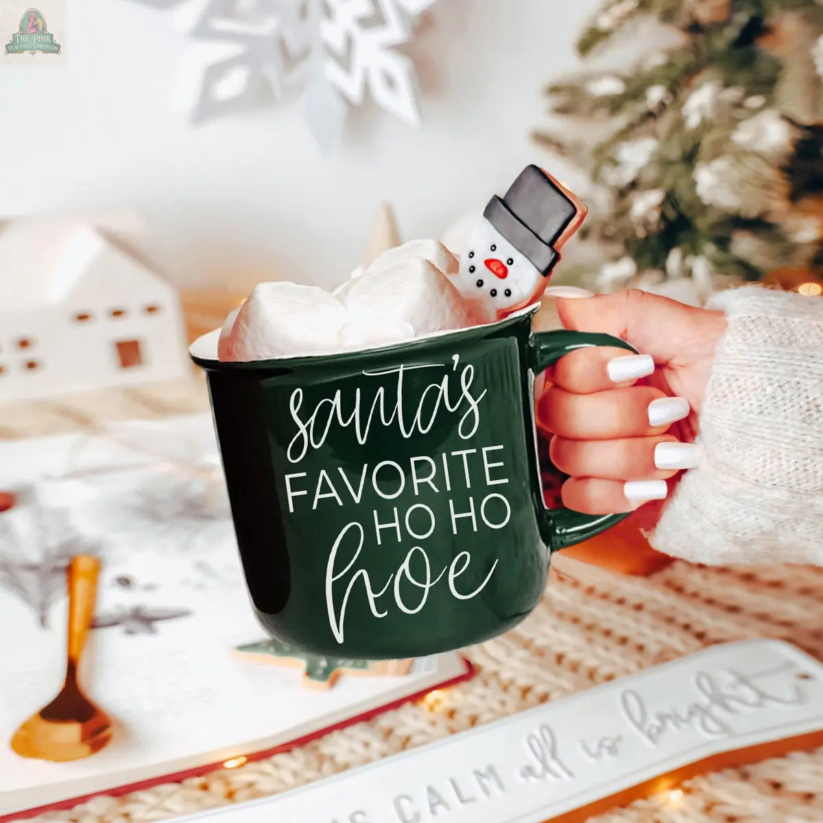 A hand with white nail polish holds the Ho Ho Hoe Mug, a green ceramic campfire mug labeled "Santas Favorite Ho Ho Hoe," filled with marshmallows and a snowman treat, set in a cozy, festive holiday scene.