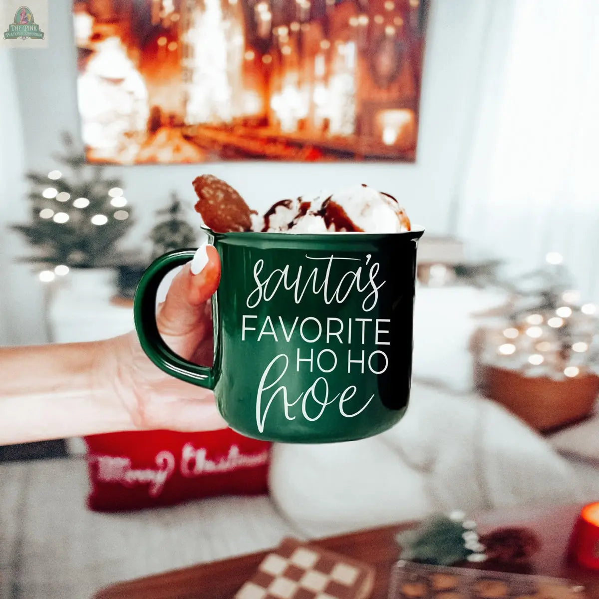 A hand holds the green ceramic Ho Ho Hoe Mug, filled with whipped cream and cookies, surrounded by festive decor, twinkling lights, and a Christmas tree in the background.