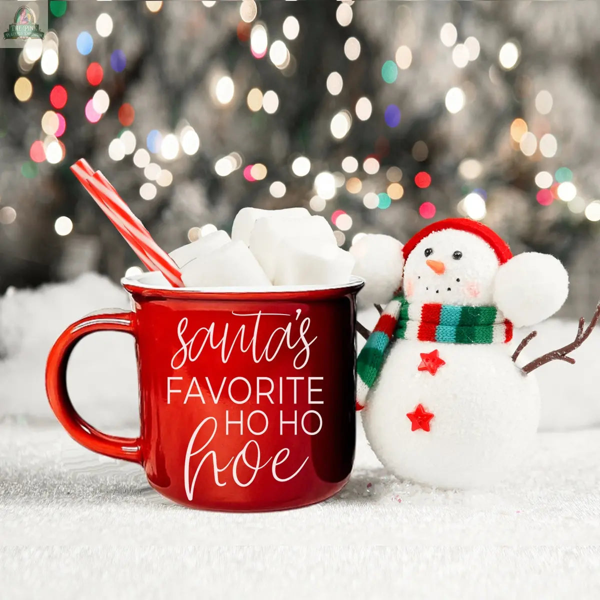 A red ceramic Ho Ho Hoe Mug filled with hot chocolate, marshmallows, and candy canes sits beside a smiling snowman on snowy ground, with festive lights twinkling in the background.