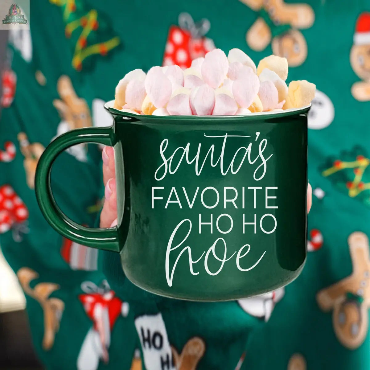 The Ho Ho Hoe Mug, a ceramic campfire-style mug featuring “Santa’s favorite ho ho hoe” in white script, is filled with marshmallows and cocoa and held by someone in a festive sweater with gingerbread men and Christmas trees.