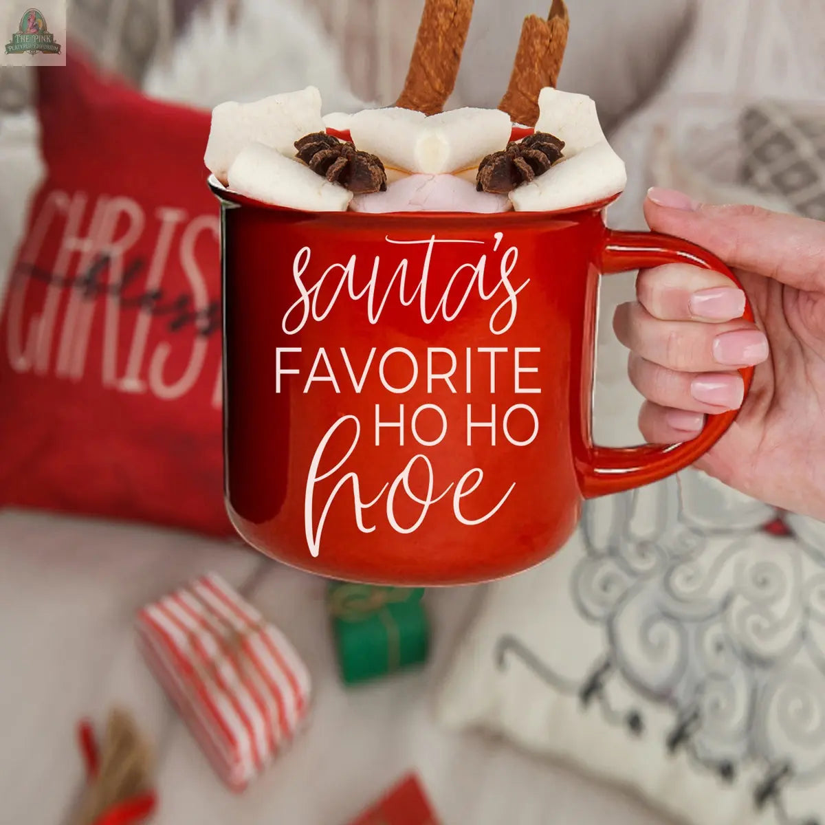 A hand holds the Ho Ho Hoe Mug, a red ceramic campfire mug filled with hot chocolate and marshmallows. “Santa’s favorite ho ho hoe” is printed on the mug, with Christmas pillows and wrapped gifts providing a cozy holiday backdrop.