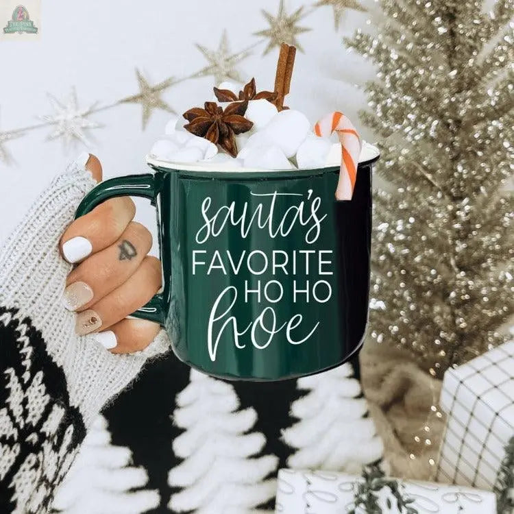 A hand holds the Ho Ho Hoe Mug filled with hot chocolate, marshmallows, cinnamon, star anise, and a candy cane. The mug features "Santa's Favorite Ho Ho Hoe" with a Christmas tree and festive decorations in the background.