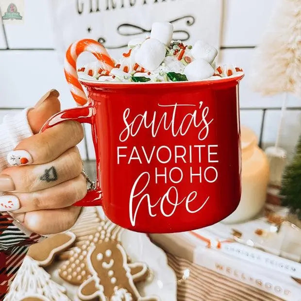 A hand with festive nail art holds the Ho Ho Hoe Mug, a ceramic campfire mug filled with whipped cream, marshmallows, and a candy cane. The mug features “Santa’s favorite ho ho hoe” in white script, surrounded by holiday decor and gingerbread cookies.