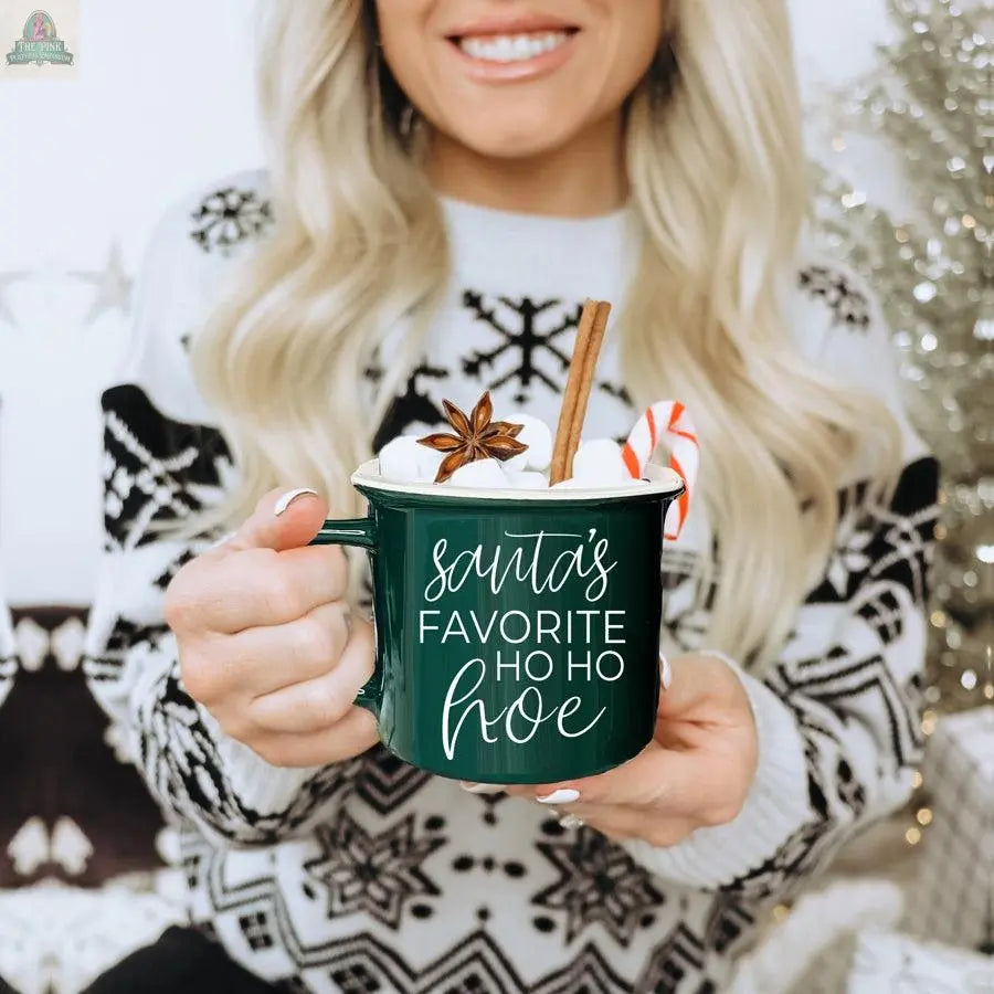 A woman in a festive sweater holds the green Ho Ho Hoe Mug filled with hot chocolate, marshmallows, cinnamon, anise, and a candy cane. The ceramic mug reads "Santa's favorite ho ho hoe" and is microwave safe—ideal for cozy nights in.