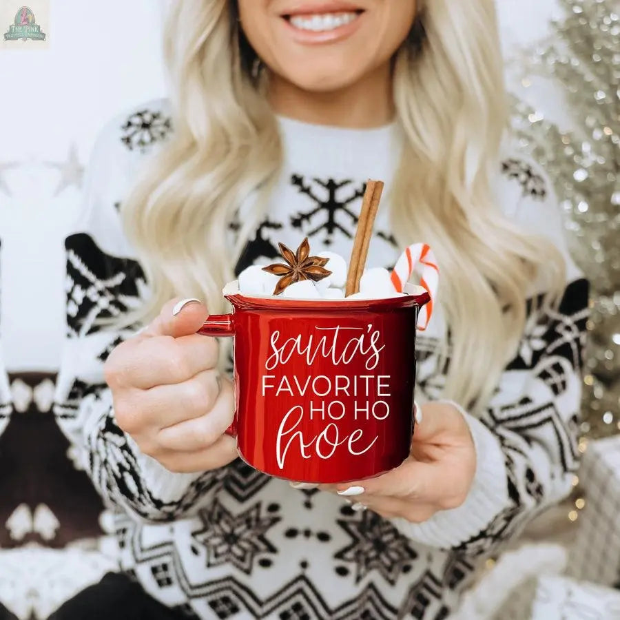 A person with long blonde hair in a black and white snowflake sweater holds the Ho Ho Hoe Mug filled with marshmallows, a candy cane, cinnamon stick, and star anise. The red ceramic mug features the text: Santa's Favorite Ho Ho Hoe.