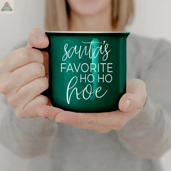 A person in a gray shirt holds the Ho Ho Hoe Mug, a green ceramic campfire cup featuring white script text that reads, "Santa's favorite ho ho hoe.
