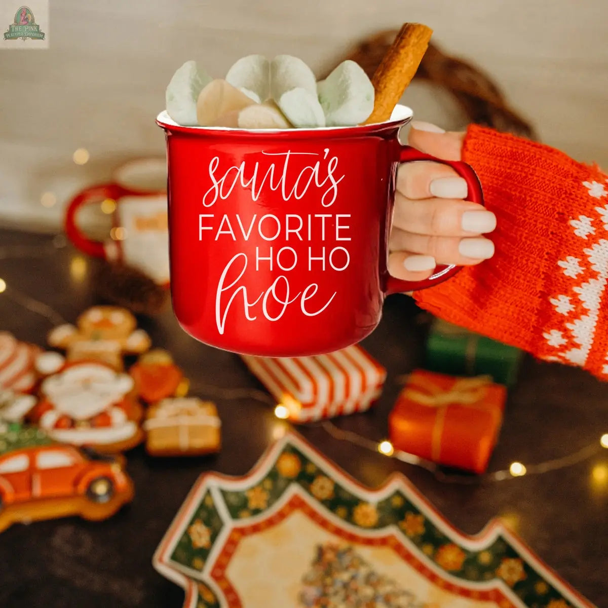 A hand in a red and white sweater holds the Ho Ho Hoe Mug with “Santa’s Favorite Ho Ho Hoe” on it, filled with marshmallows and a cinnamon stick. Christmas cookies, gifts, and string lights blur in the festive background.
