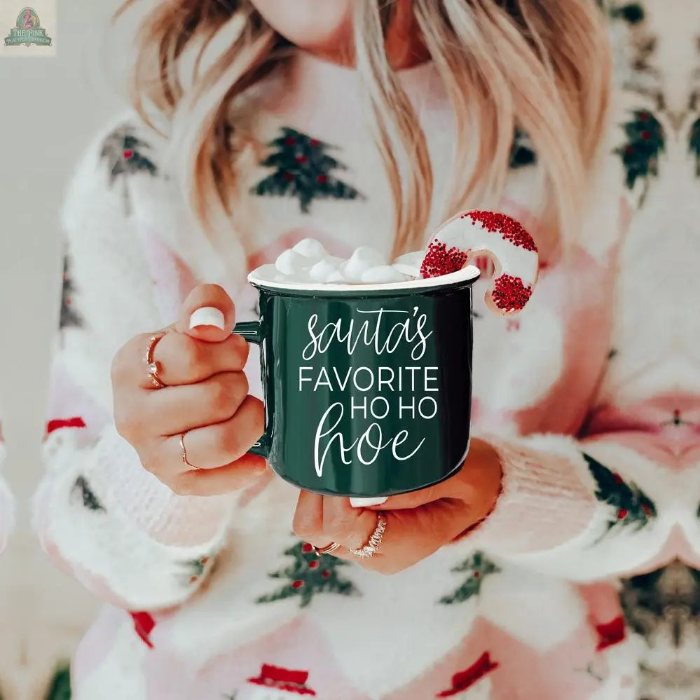 A woman in a festive sweater holds the Ho Ho Hoe Mug, a green ceramic campfire-style mug reading “Santa’s Favorite Ho Ho Hoe.” Topped with marshmallows and a candy cane, this microwave safe mug is perfect for holiday cheer.