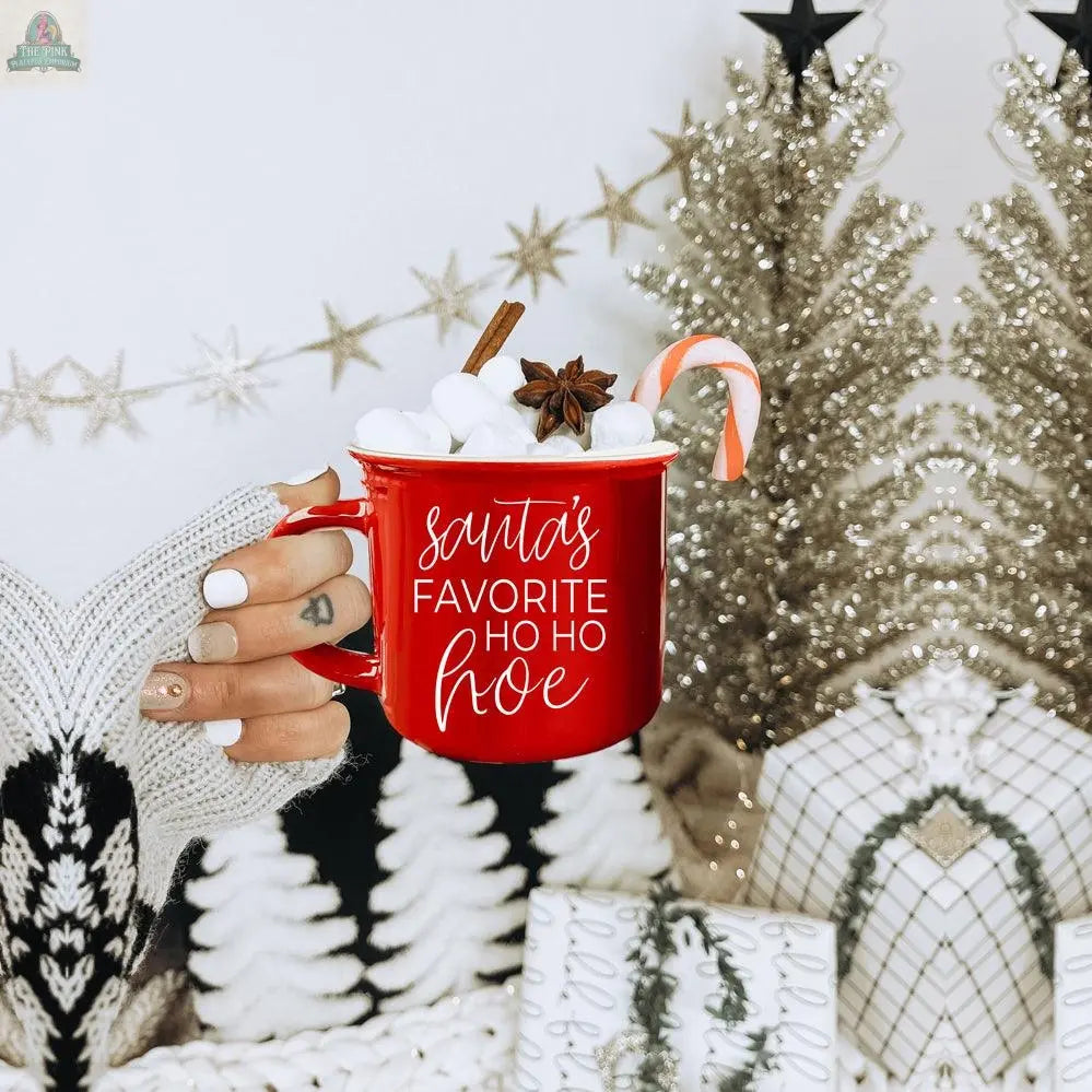 A hand wearing black and white knit gloves holds the Ho Ho Hoe Mug—a red ceramic campfire mug filled with marshmallows, a candy cane, cinnamon stick, and star anise. Christmas trees and wrapped gifts decorate the background.