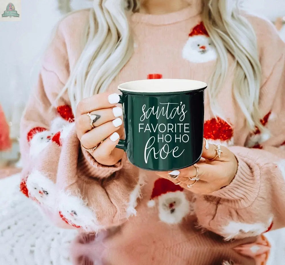 A woman with long blonde hair and white nails, wearing a pink Santa-themed sweater, holds the Ho Ho Hoe Mug—a dark green handmade ceramic campfire mug featuring “Santa’s favorite HO HO HOE” in white script.