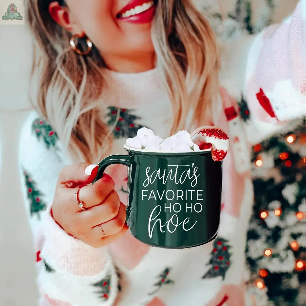 A woman in a festive sweater holds the Ho Ho Hoe Mug, a green ceramic campfire mug with “Santa’s Favorite Ho Ho Hoe” printed on it, topped with marshmallows and a candy cane, in front of a decorated Christmas tree.