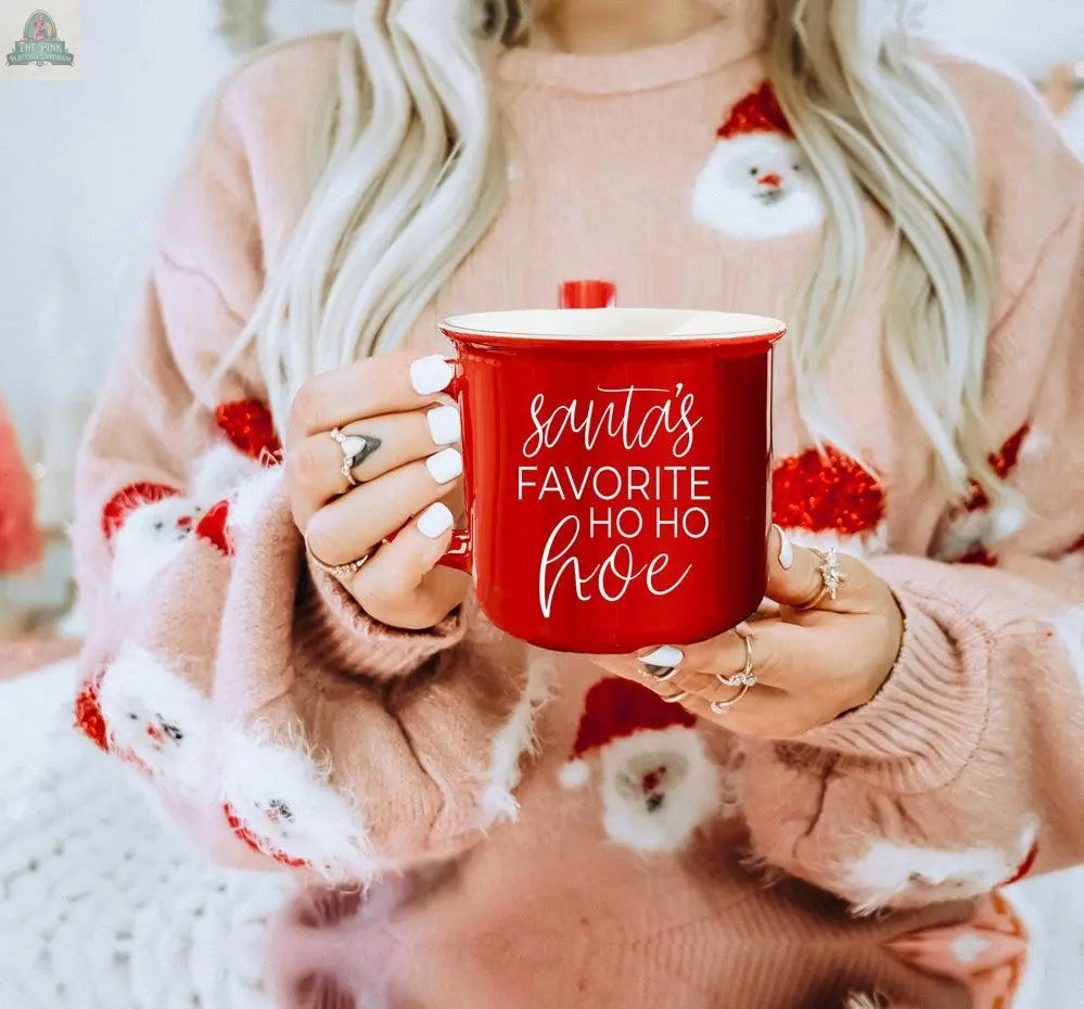 A woman with long blonde hair in a Santa sweater holds the Ho Ho Hoe Mug, a red ceramic campfire mug featuring "Santa’s favorite ho ho hoe" in white script.