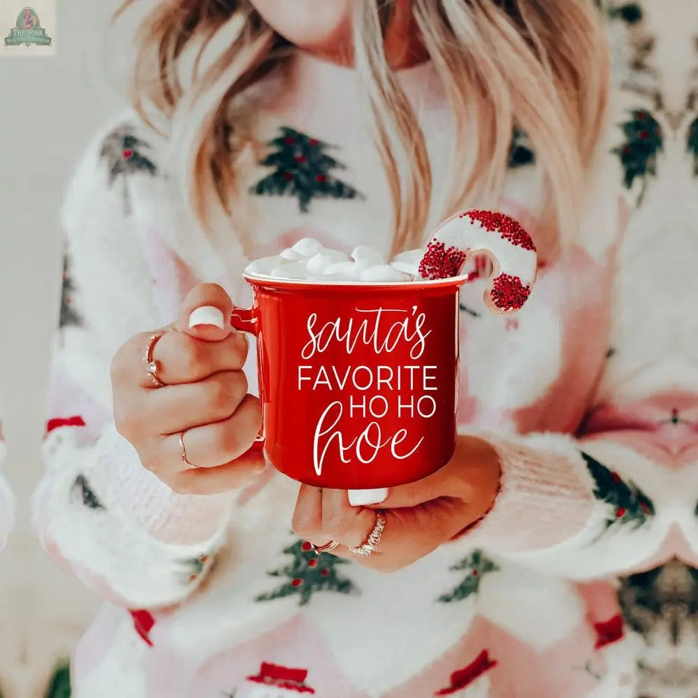A person in a festive sweater holds the microwave-safe Ho Ho Hoe Mug, a red ceramic campfire mug filled with marshmallows and a candy cane, featuring "Santa’s Favorite Ho Ho Hoe" in playful white script.