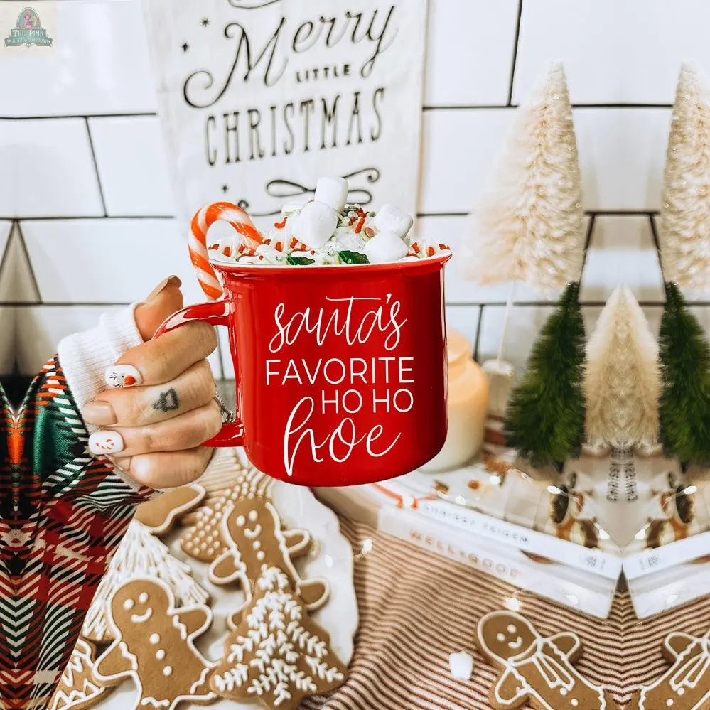 A hand holds a red Ho Ho Hoe Mug filled with marshmallows in front of Christmas decorations and a "Merry little Christmas" sign. A decorated gingerbread cookie is also visible.
