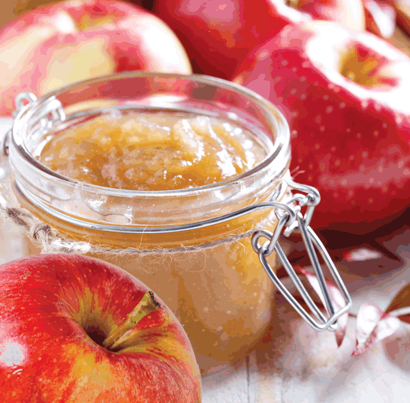 An open glass jar of smooth, light brown Hot Apple Butter Atomizer Oil sits on a table with whole red apples, filling the air with a comforting, warm fragrance.