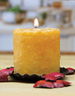 The Hot Apple Butter Mini Hearth candle by Pink Platypus Emporium, with its textured yellow design, glows on a black holder amid red dried petals, set against a gently blurred backdrop.