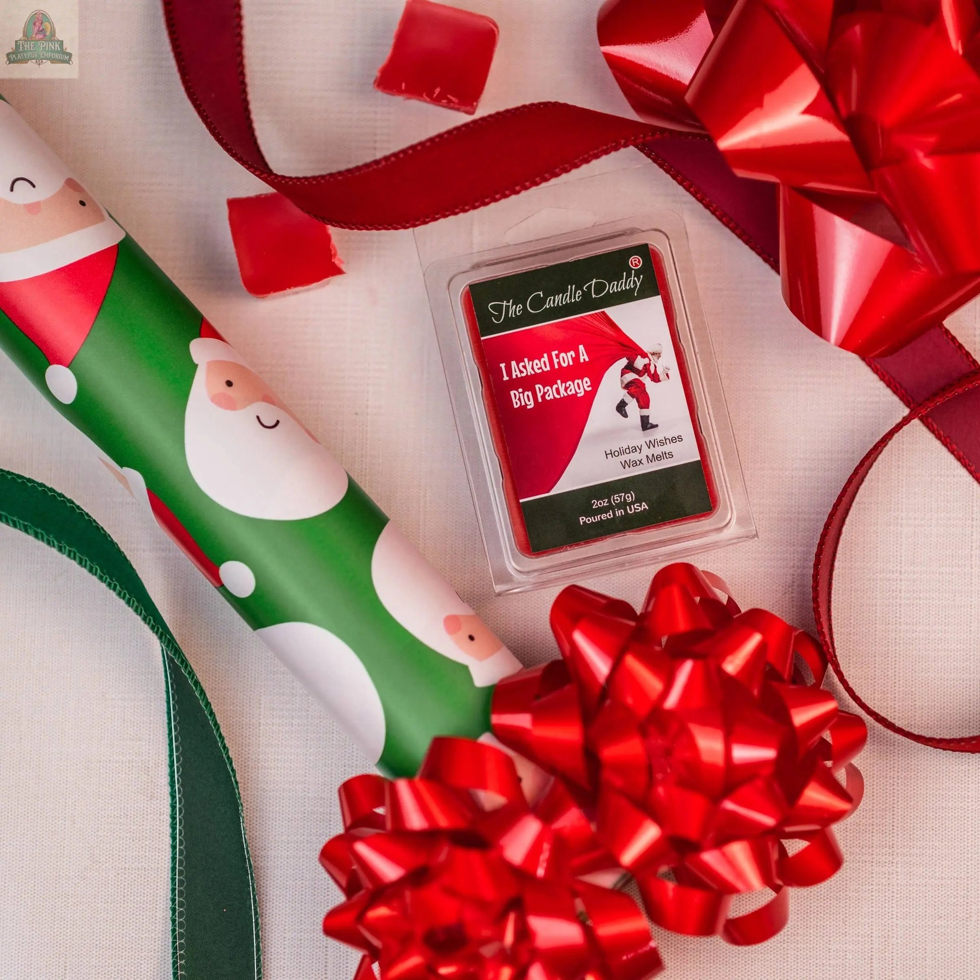 A festive flat lay with Santa wrapping paper, big red bows, and I ASKED FOR A BIG PACKAGE - HOLIDAY WISHES SCENTED WAX MELTS arranged on a white surface.