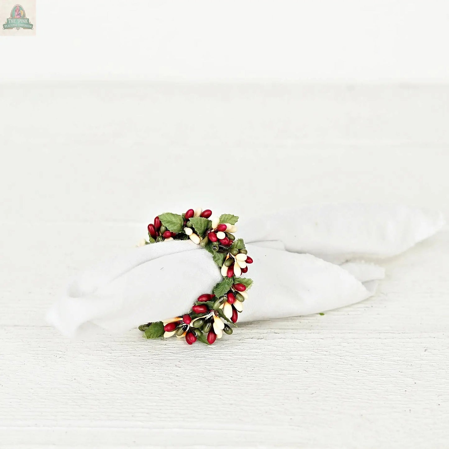 The ISB13700FH 1.5in Pip Berry Napkin Ring-Farmhouse holds a rolled white cloth napkin, featuring red, white, and green artificial berries and leaves for a charming accent on a white wooden surface.