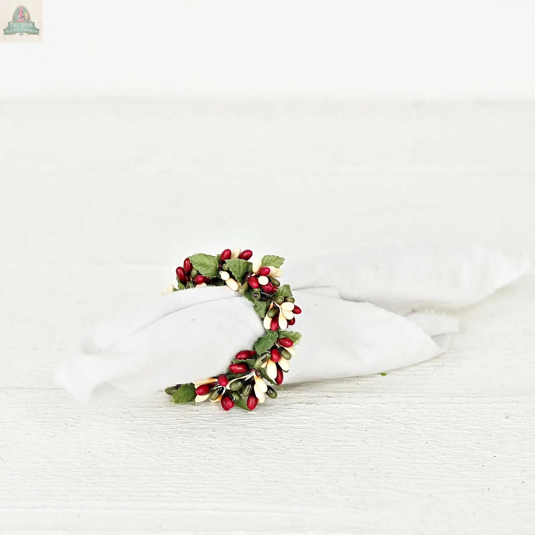 The ISB13700FH 1.5in Pip Berry Napkin Ring-Farmhouse holds a rolled white cloth napkin, featuring red, white, and green artificial berries and leaves for a charming accent on a white wooden surface.