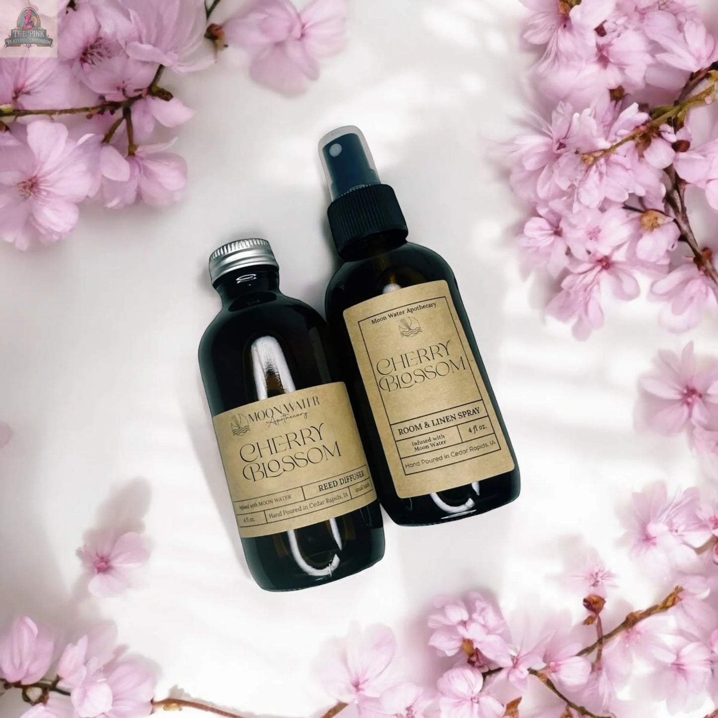 Two dark glass bottles labeled “Moon Water Apothecary: Japanese Cherry Blossom - ROOM SPRAY” rest on a white surface with pink sakura flowers, creating a softly lit scene and evoking a calming floral aroma.