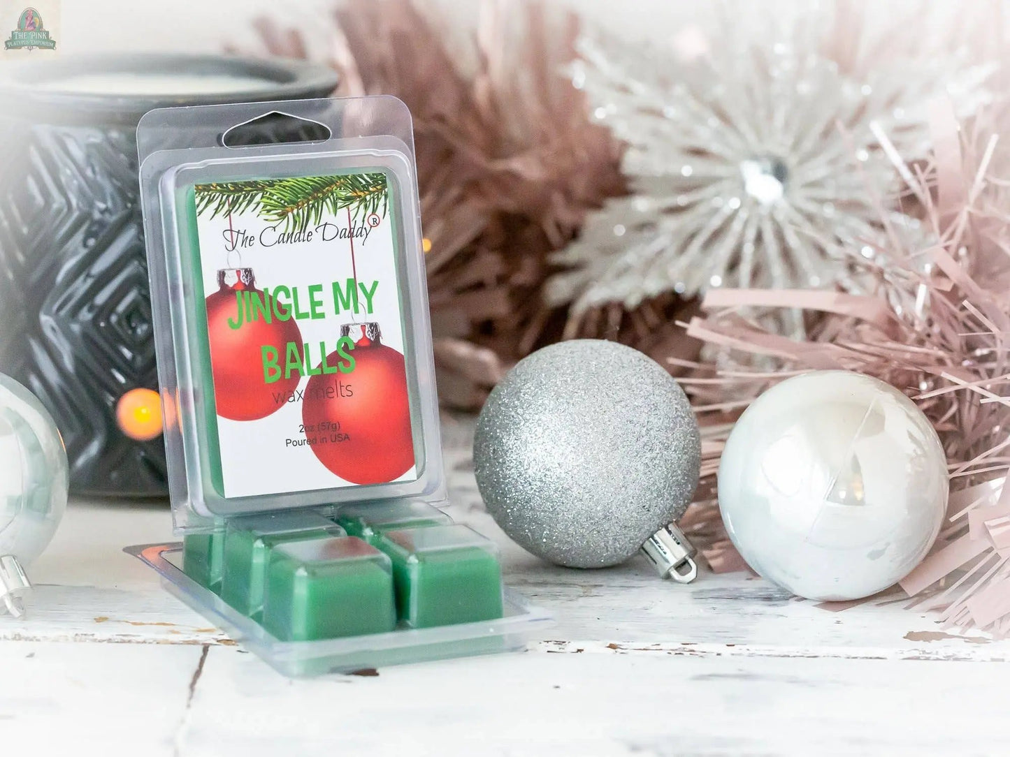 A pack of Jingle My Balls - Holly Berry Funny Christmas Scent wax melts rests on a white surface amid silver and white Christmas ornaments and pink tinsel, with a black wax warmer visible in the background.