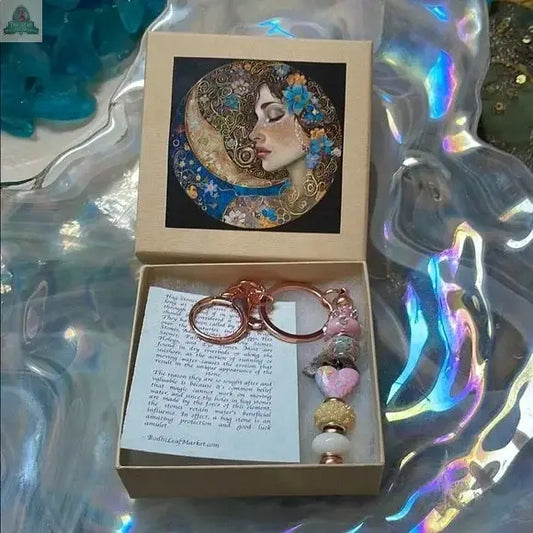 The Lady of The Moon Gift Box with Hagstone Beaded Keychain features an artistic woman-in-moon design, a pink and gold beaded charm, and a note, all on an iridescent wavy surface—ideal for Sanibel Island keepsake lovers.