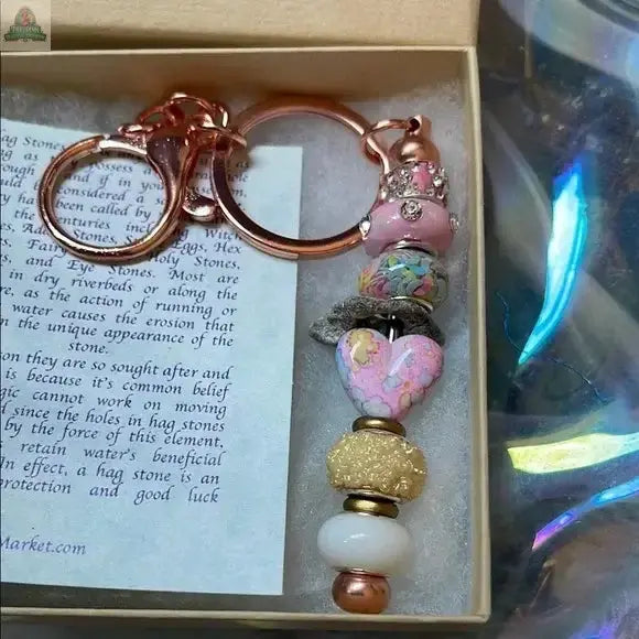 The Lady of The Moon Gift Box features a rose gold keychain with pink, gold, white, and marbled beads. Presented in a small box on a printed note, it rests against a soft, iridescent background inspired by Sanibel Island.
