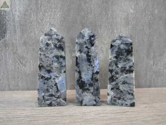 Three Larvikite Flat Obelisks from Keystone Crystals—shimmering grounding crystal towers with blue, black, and grey speckles—stand upright on wood, perfect for energy work. Available at Pink Platypus Emporium against a neutral textured background.
